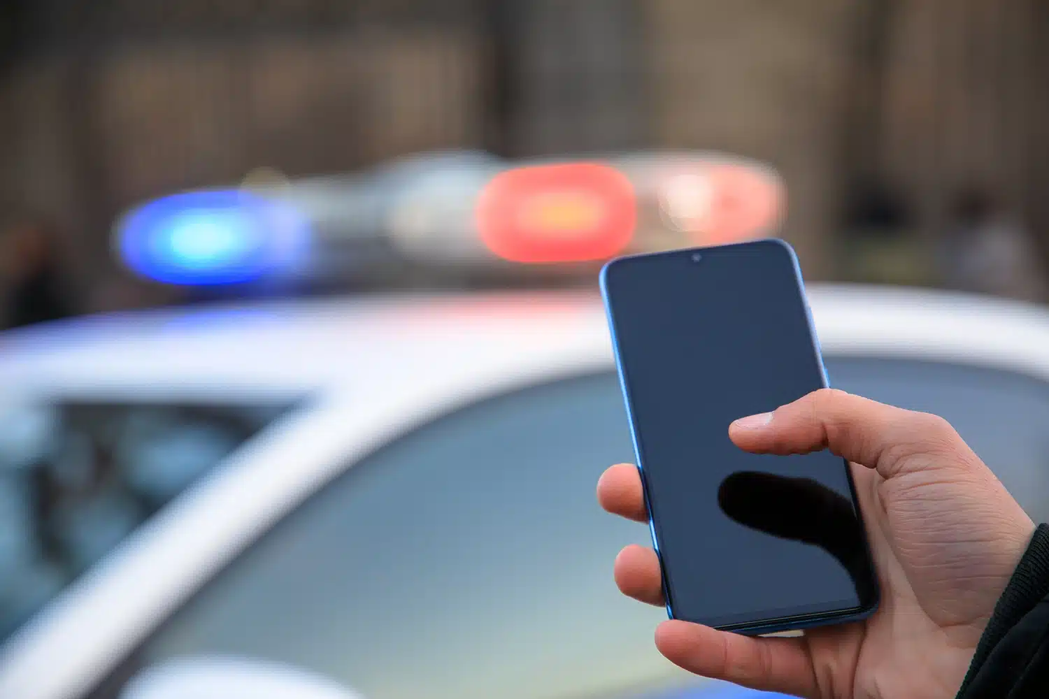 Police officer holding a cell phone with police cruiser lights in the background